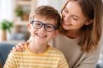 gyermek szemüvegben mosolyog, büszke anya ölelésében